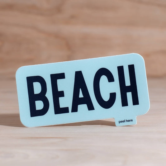 A light blue rectangular sticker with rounded corners and the word 'BEACH' printed in bold black letters, shown standing on a light wood surface with a blurred wood background. A small 'peel here' label is visible on the bottom right of the sticker.