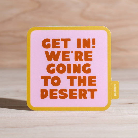 A square pink sticker with rounded corners, featuring the text 'GET IN! WE'RE GOING TO THE DESERT' in orange lettering, with a yellow border and a small side tab that reads 'post it'. The sticker is displayed on a light wood surface.