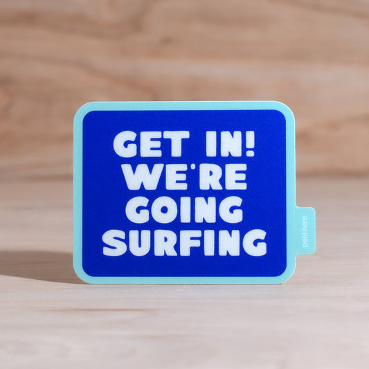 A square blue bumper sticker with rounded corners and a light blue border, featuring the bold white text 'GET IN! WE'RE GOING SURFING'. The sticker is shown standing upright on a light-colored wooden surface with a matching wooden background.