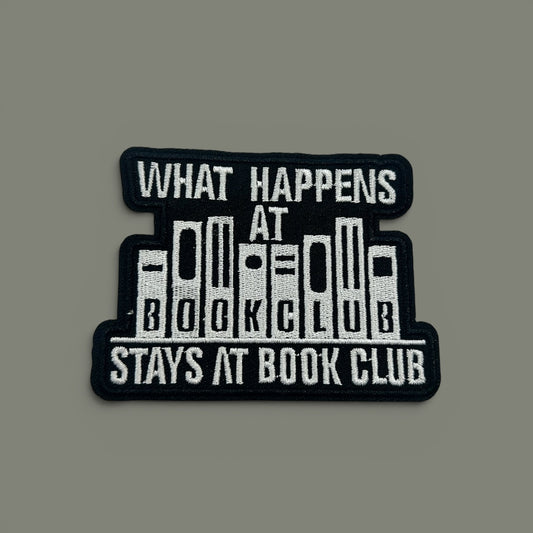 A rectangular embroidered patch with a black background and white text reading 'WHAT HAPPENS AT BOOK CLUB STAYS AT BOOK CLUB,' with stylized book spines forming the center, shown on a plain gray background.