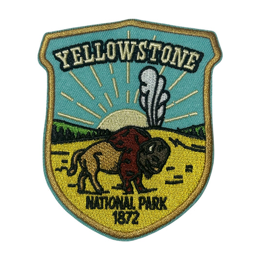 An embroidered shield-shaped patch for Yellowstone National Park, featuring a bison, a puff of smoke, a yellow sun, green trees, and the text 'YELLOWSTONE NATIONAL PARK 1872' on a white background.