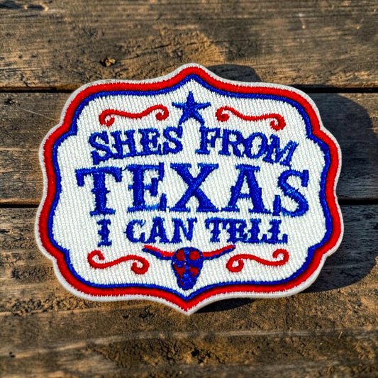 An embroidered patch with the text 'SHES FROM TEXAS I CAN TRUST' in blue lettering on a white background, bordered in red and blue, placed on a rustic wooden surface.