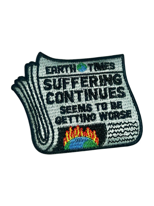 An embroidered patch shaped like a folded newspaper with the text 'EARTH TIMES SUFFERING CONTINUES SEEMS TO BE GETTING WORSE' and a small multicolor globe-and-flames graphic near the bottom, shown on a plain white background.