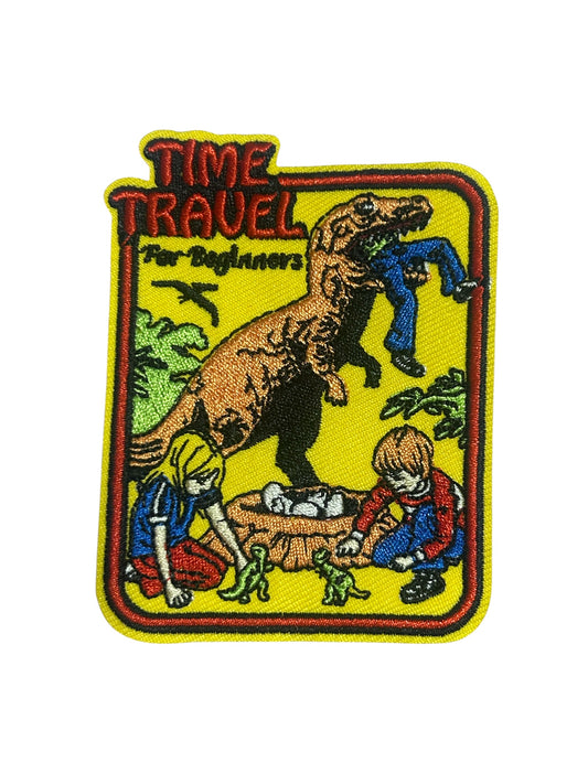 An embroidered patch with a yellow background and the text 'TIME TRAVEL For Beginners' at the top. The patch features a brown dinosaur carrying a person in its mouth, with children and small dinosaurs below, all outlined in red and yellow.