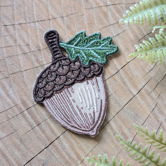 Embroidered acorn patch on a wooden surface with green leaves.