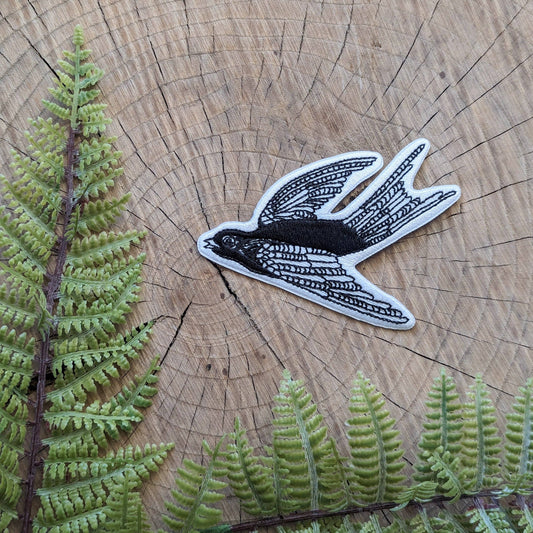A black and white embroidered patch of a flying swallow bird, styled on a rustic wood slice background with green fern leaves.