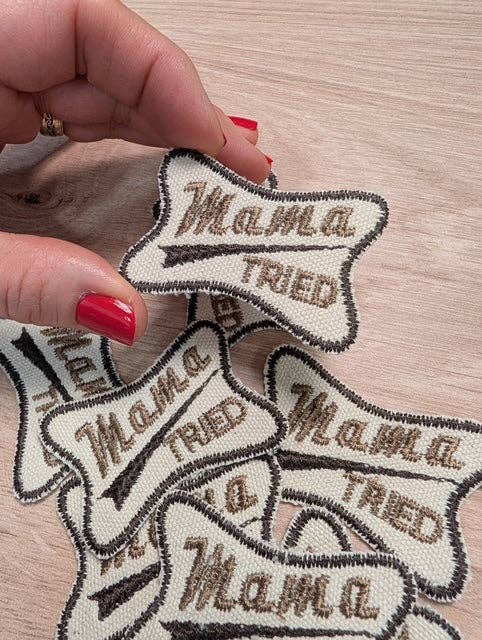 Hand holding a 'Mama Tried' patch with multiple patches on a wooden surface