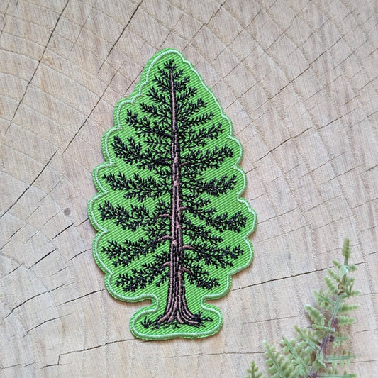 A green embroidered pine tree patch with black and brown stitching, displayed on a natural wood slice background.