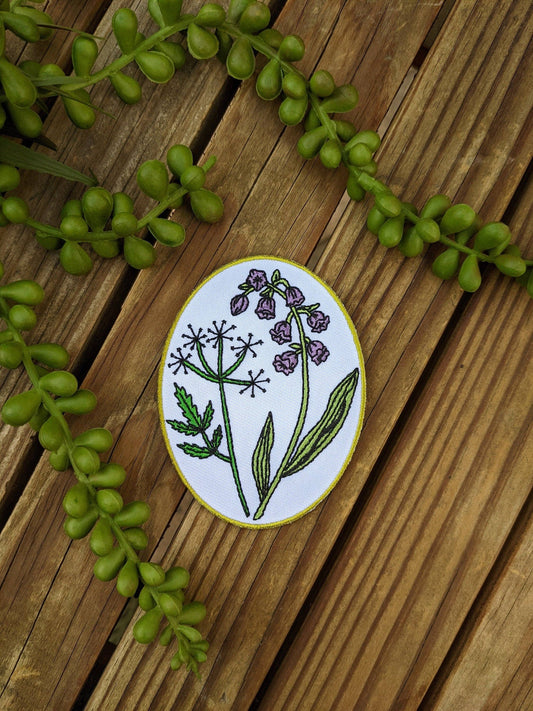 An oval enamel piatch featuring a botanical illustration of purple flowers and green leaves on a white background, bordered in gold. The pin is displayed on a rustic wooden surface decorated with green beaded vines.
