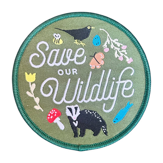 A round embroidered patch with a green border and olive green background, featuring the words “Save Our Wildlife” in white script, surrounded by small illustrated animals and nature motifs including a bird, bee, flower, butterfly, badger, mushroom, and fish.