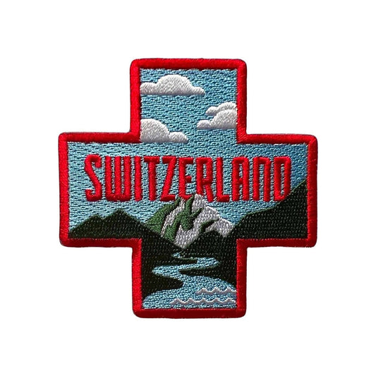 An embroidered cross-shaped patch featuring a Switzerland landscape with red border, red 'SWITZERLAND' text, blue sky, white clouds, green and gray mountains, and a blue river on a white background.