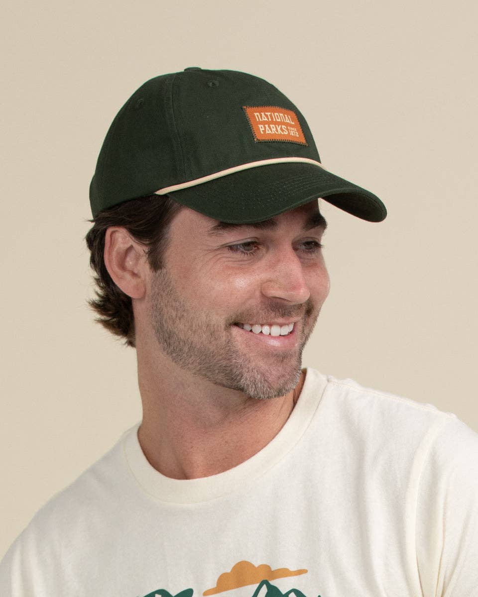 A man wearing a dark green baseball cap with a tan patch that reads 'NATIONAL PARKS NET' and a cream t-shirt, photographed against a solid beige background.