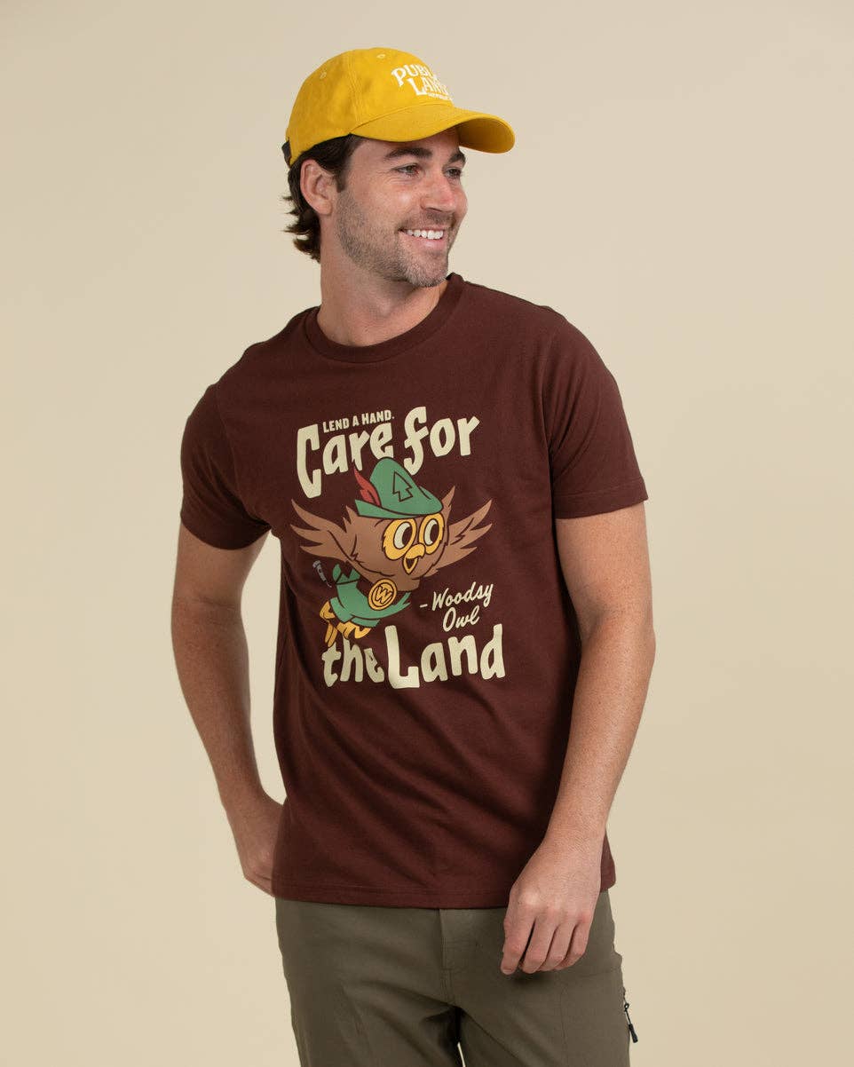 A man wearing a brown graphic t-shirt with a cartoon owl design and a yellow baseball cap, standing against a solid beige background.