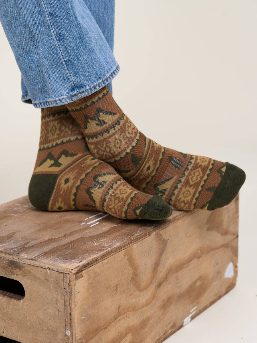 A pair of patterned crew socks worn with blue jeans, resting on a rustic wooden crate against a plain off-white background.