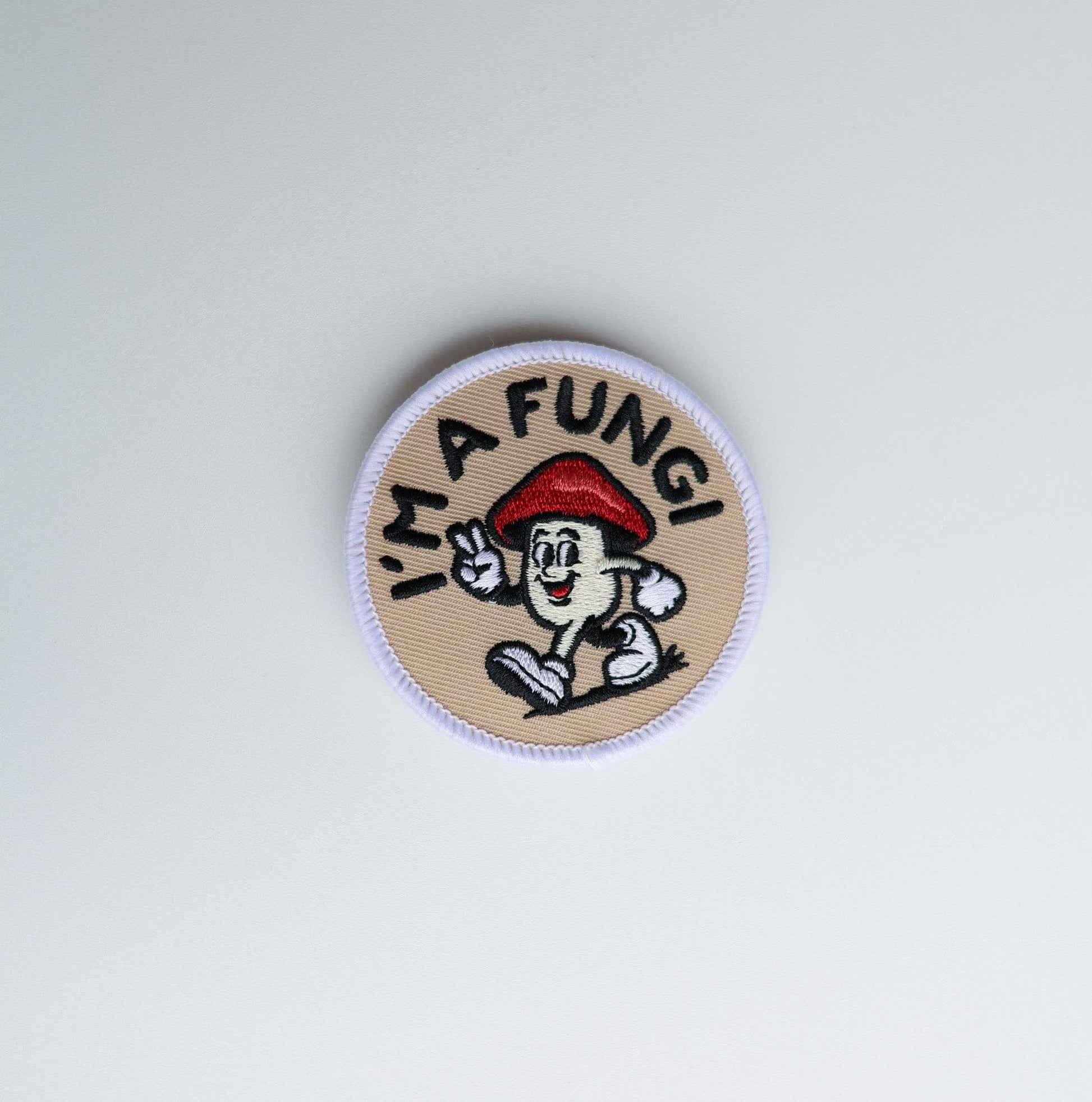 A circular embroidered patch featuring a cartoon mushroom character wearing a red beret and waving, with the text 'I'M A FUNGI' in black around the character. The patch has a beige background with a white stitched border and is photographed on a plain light gray background.
