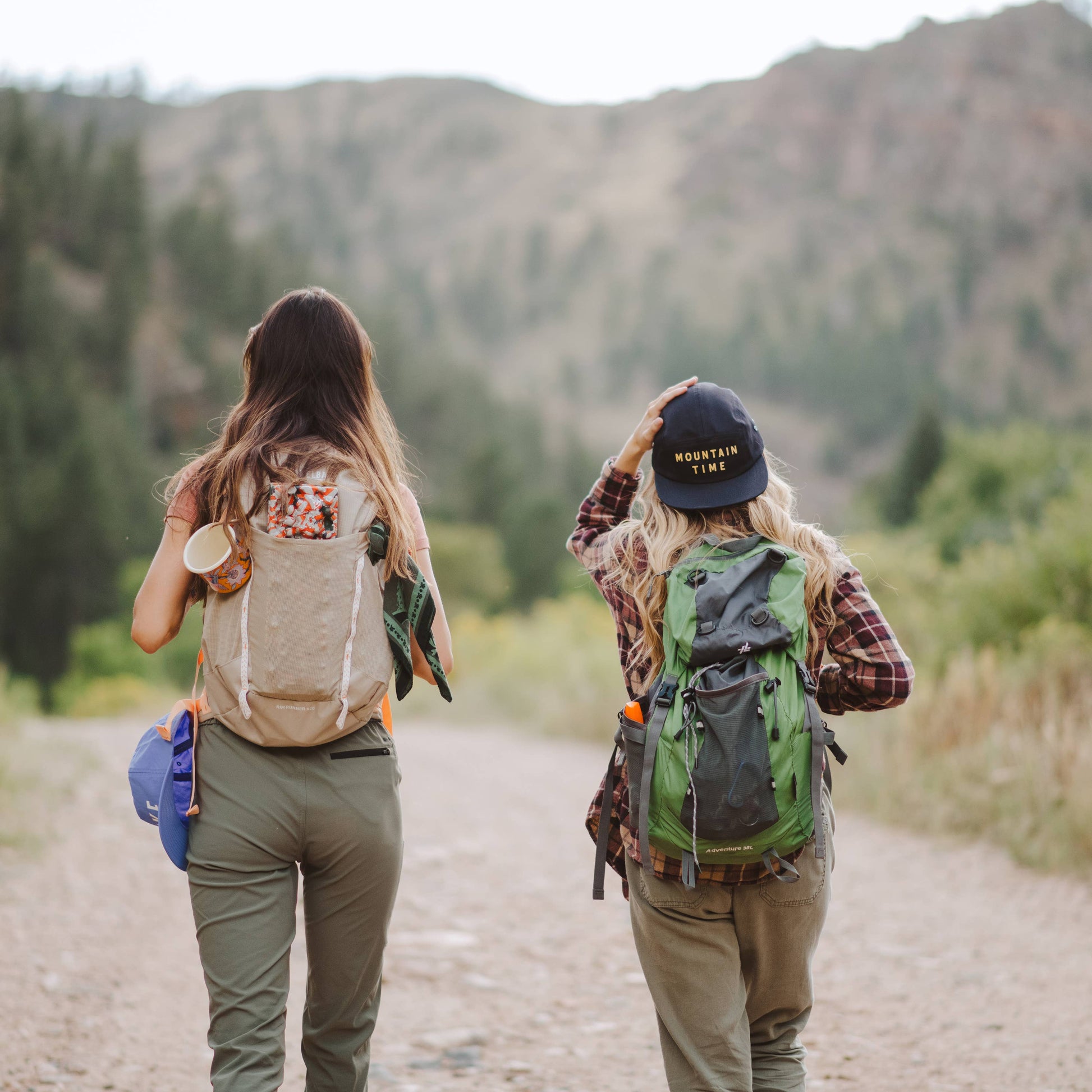 Two women hiking on a dirt path with mountains in the background, shown from behind. One wears a beige backpack and the other wears a green and black backpack, with one holding a blue item.