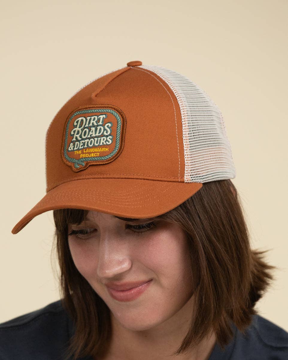 A woman wearing a rust-orange and white mesh trucker hat with a 'Dirt Roads & Detours' patch on the front, photographed against a solid beige background.
