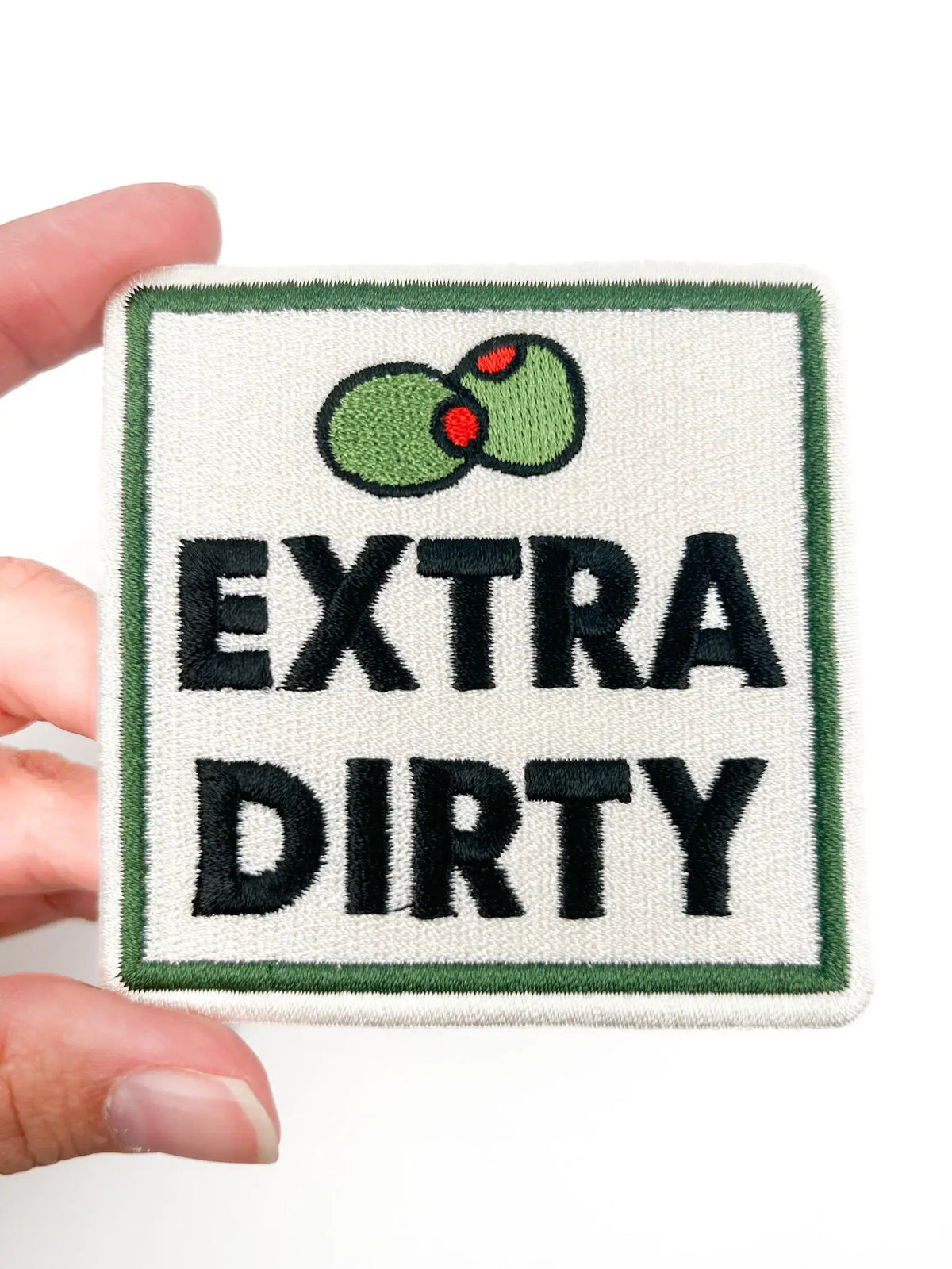 Patch with 'Extra Dirty' text and olives held by a hand on a white background