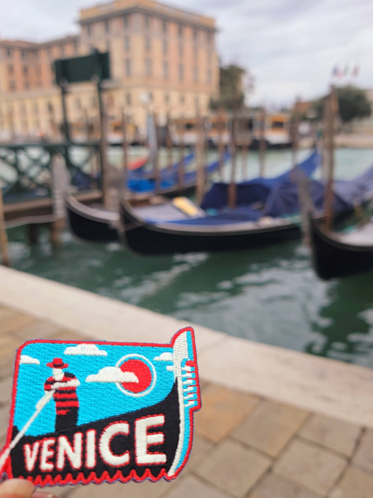 A close-up of a Venice embroidered patch in the foreground with a blurred background showing gondolas in a canal and a building.