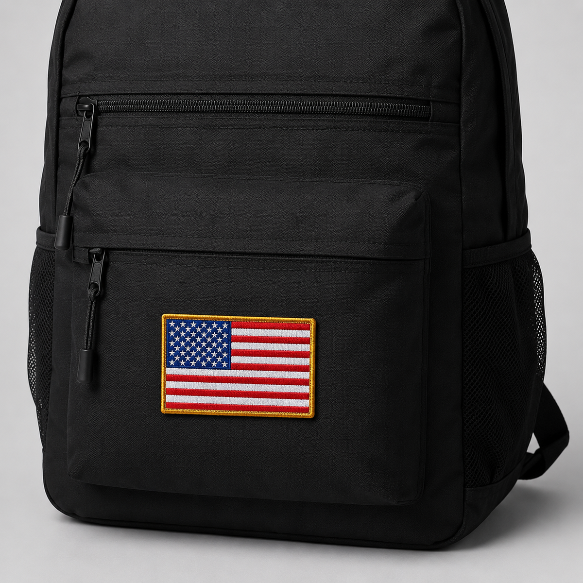 A black backpack with a rectangular embroidered American flag patch on the front pocket, shown against a light gray background.
