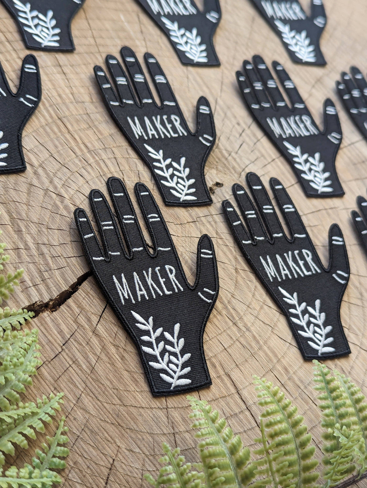 Black hand-shaped patches with 'MAKER' text on a wooden surface with ferns.