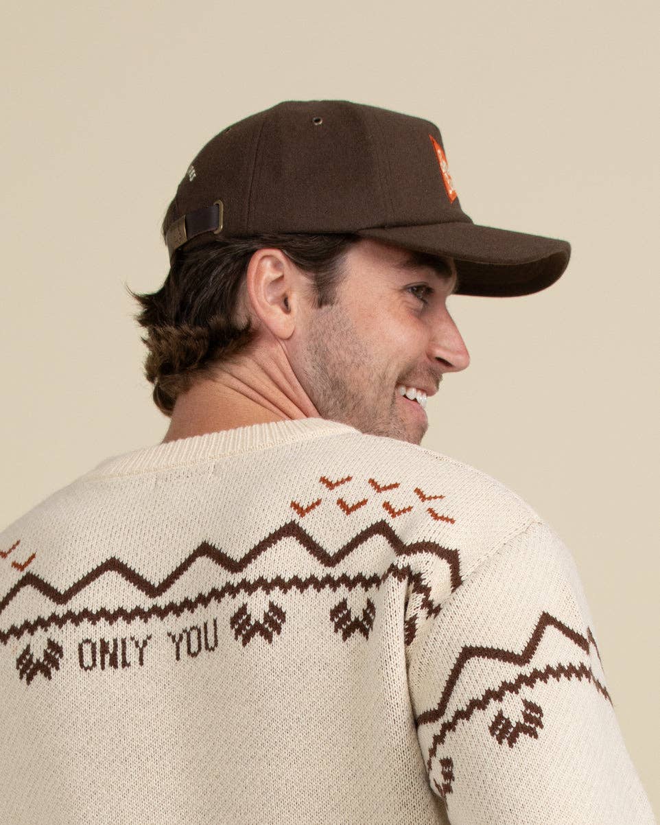 A man wearing a brown baseball cap and a cream knit sweater with brown geometric patterns and the text 'ONLY YOU' on the back, shown from a side-back angle against a plain beige background.