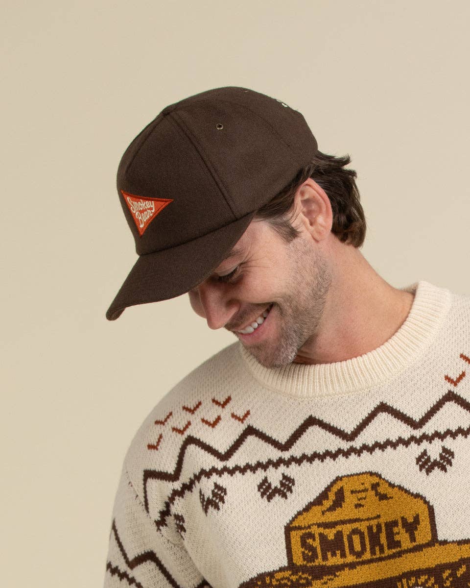 A man wearing a dark brown baseball cap with a small orange embroidered logo and a cream knit sweater with a brown patterned design and a yellow 'SMOKEY' graphic, set against a solid beige background.