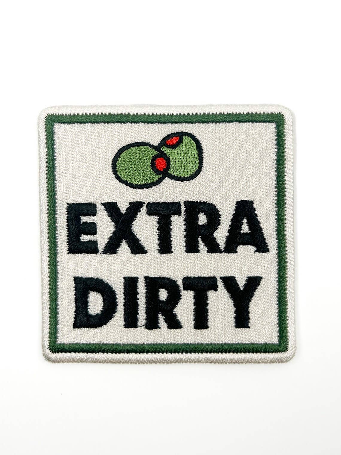 An embroidered square patch with a white background, green border, black text reading 'EXTRA DIRTY', and two green olives with red pits near the top.