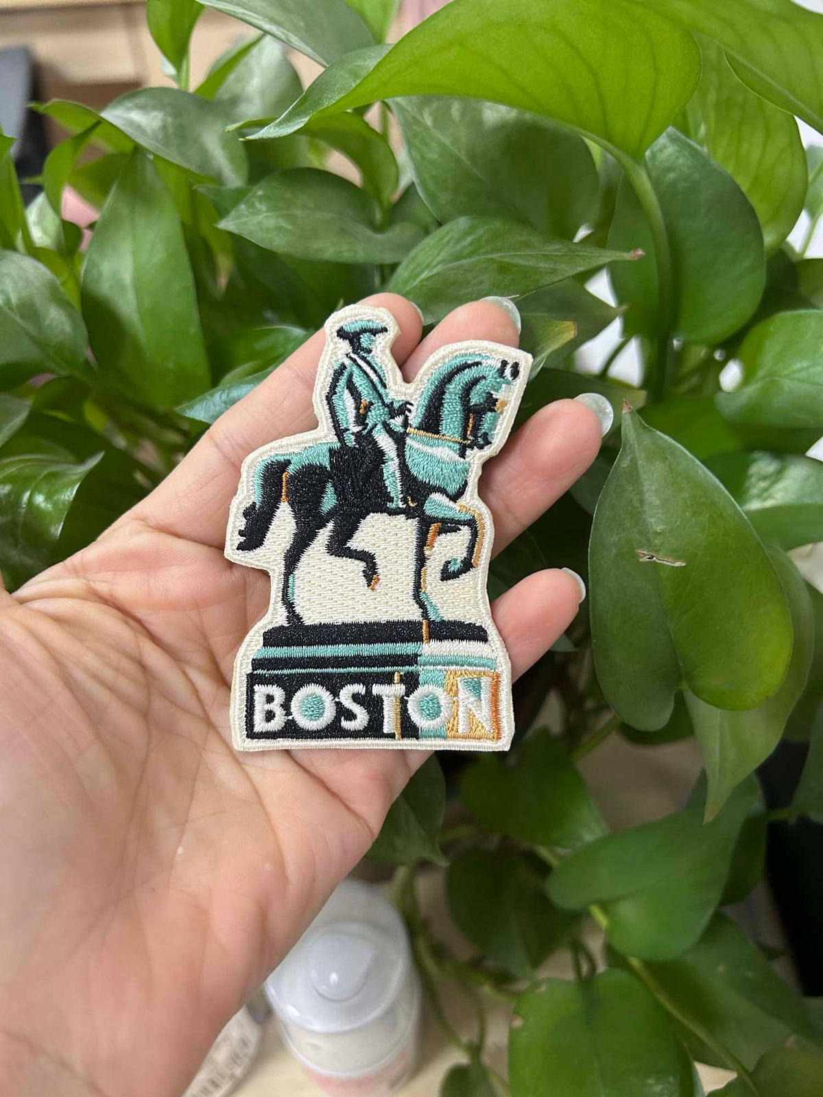 Hand holding a patch with a horse and rider design labeled 'Boston' against a green leafy background.