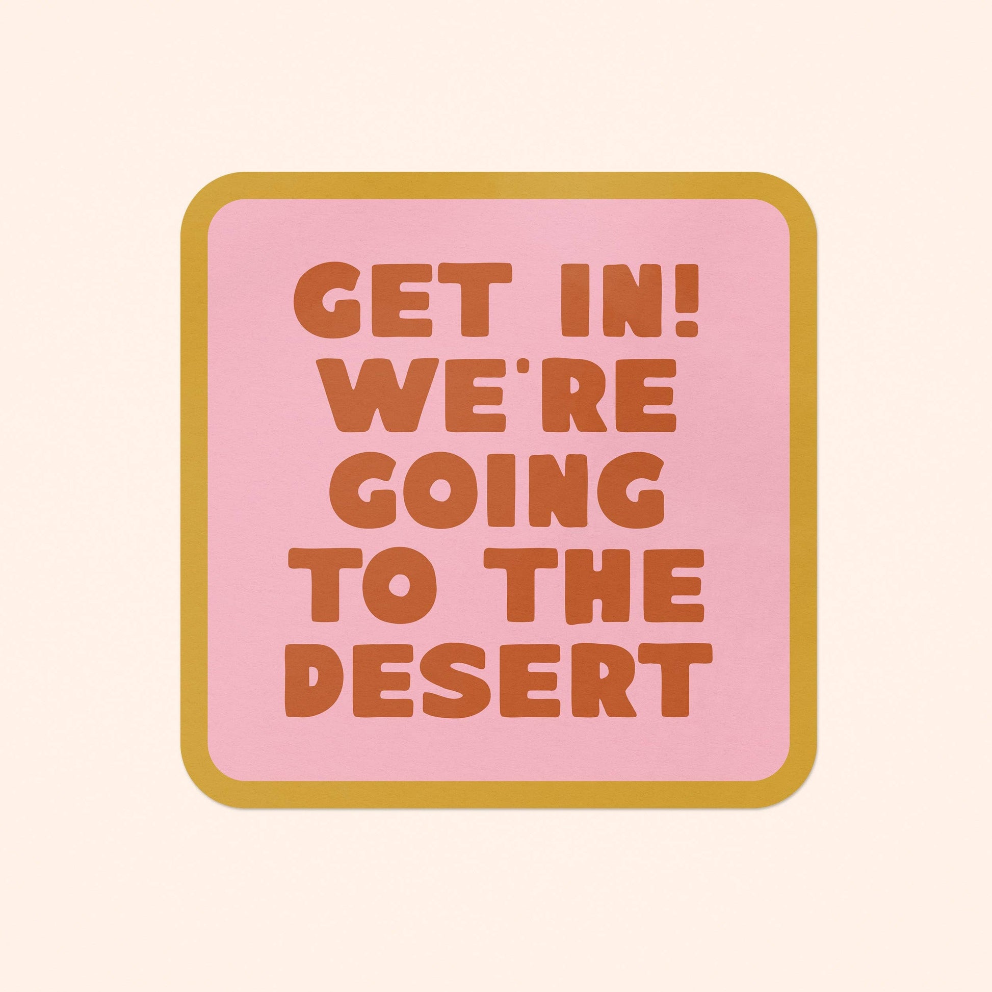 A square pink sign or coaster with rounded corners, outlined in mustard yellow, featuring the text 'GET IN! WE'RE GOING TO THE DESERT' in orange-brown block letters on a light cream background.