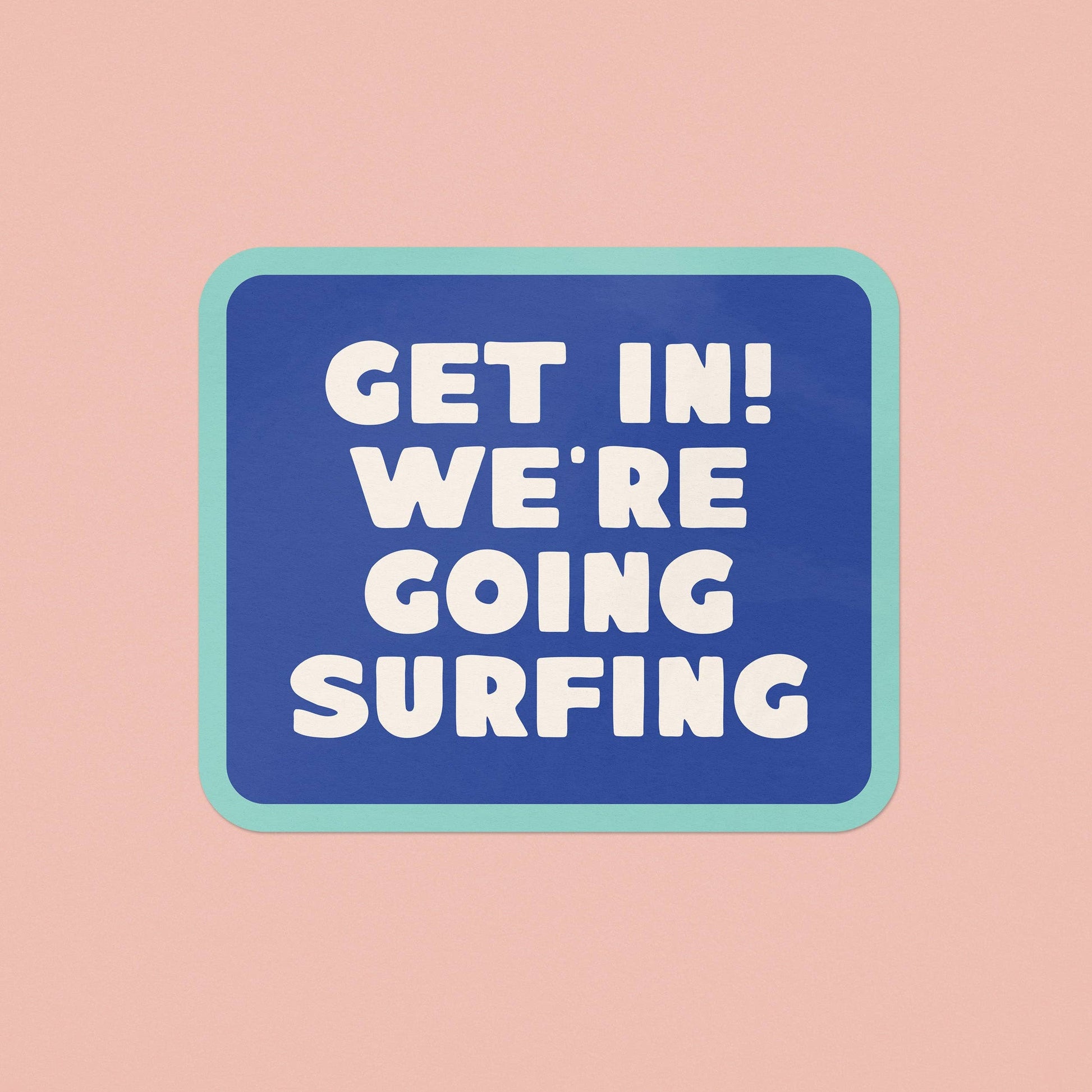 A square graphic sticker or sign with rounded corners featuring the text 'GET IN! WE'RE GOING SURFING' in bold white letters on a blue background, bordered by a light teal outline, centered on a solid pale pink background.
