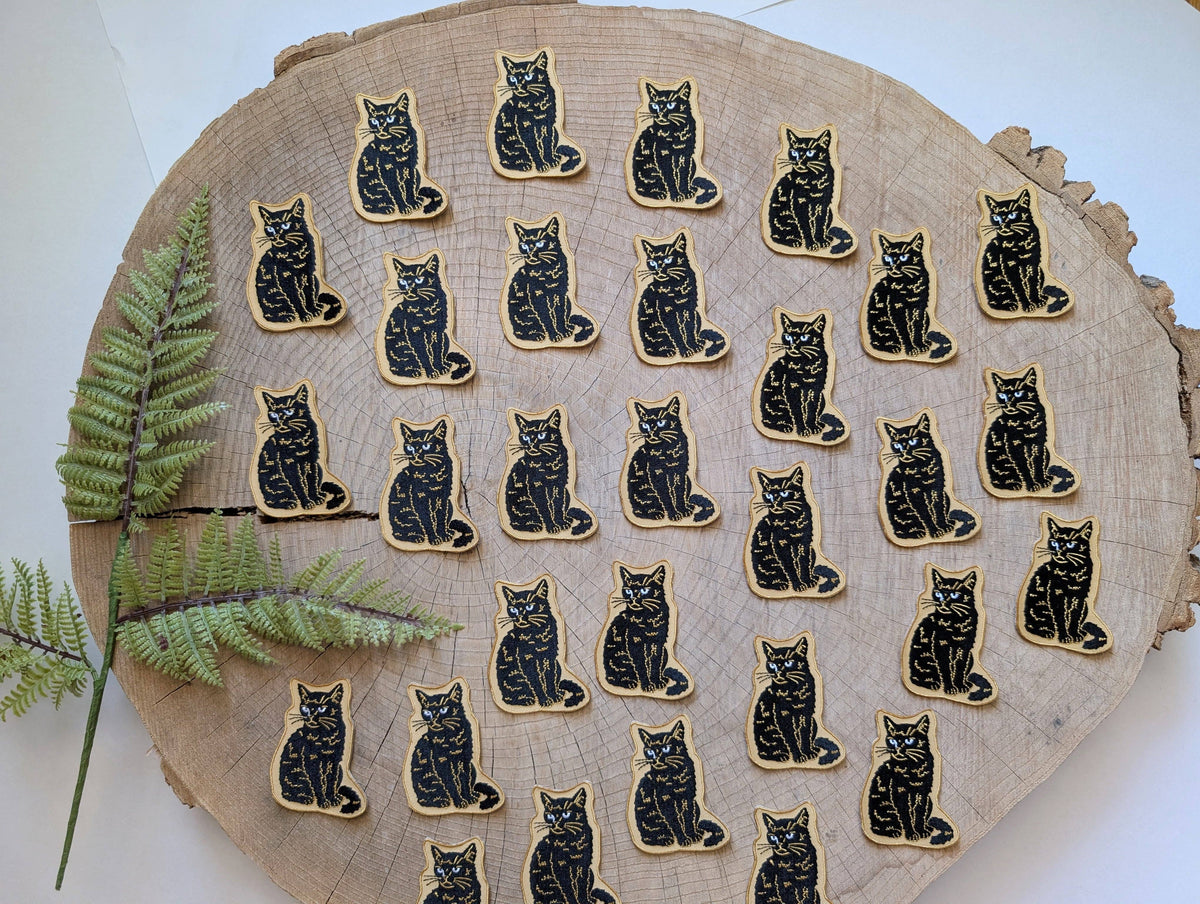 Black cat illustrations on a wooden surface with a fern leaf.