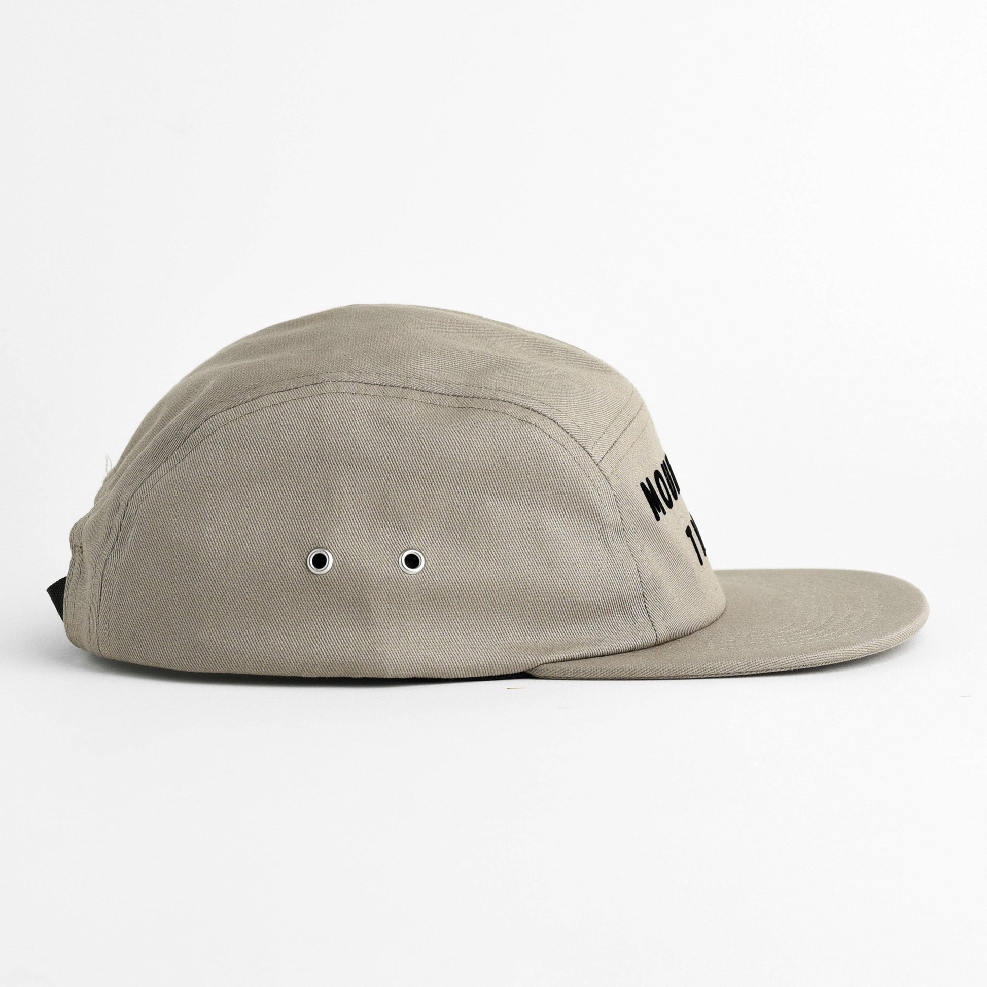 A light beige 5-panel camp cap with a flat brim, featuring black embroidered text on the side and ventilation eyelets, photographed on a clean white background.