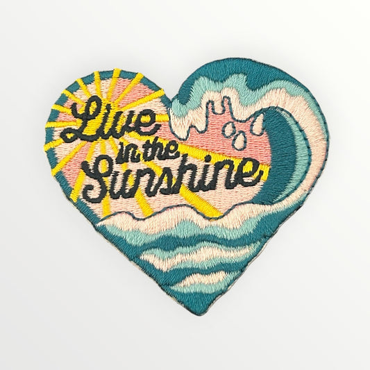 Heart-shaped embroidered patch featuring a stylized ocean wave and sunburst with the text 'Live in the Sunshine' in black script, shown on a plain white background.