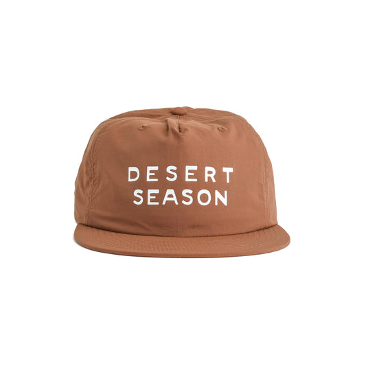 A brown flat-brim quick dry cap with the words 'DESERT SEASON' printed in white block letters on the front, shown against a plain white background.