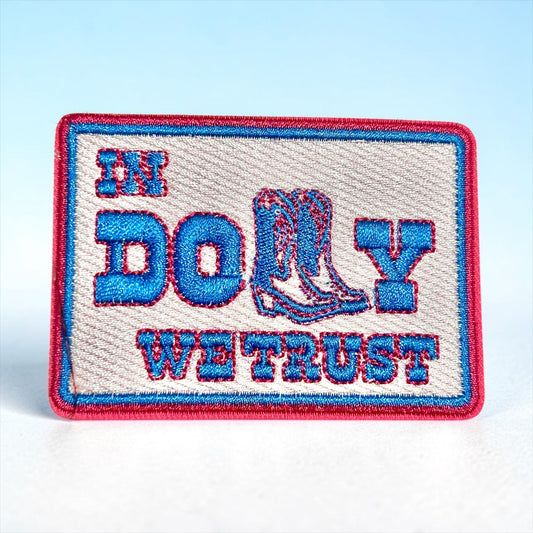 An embroidered rectangular patch with the text 'IN DOLLY WE TRUST' in blue and red, featuring a cowboy boot motif, on a light beige fabric background with a red border.