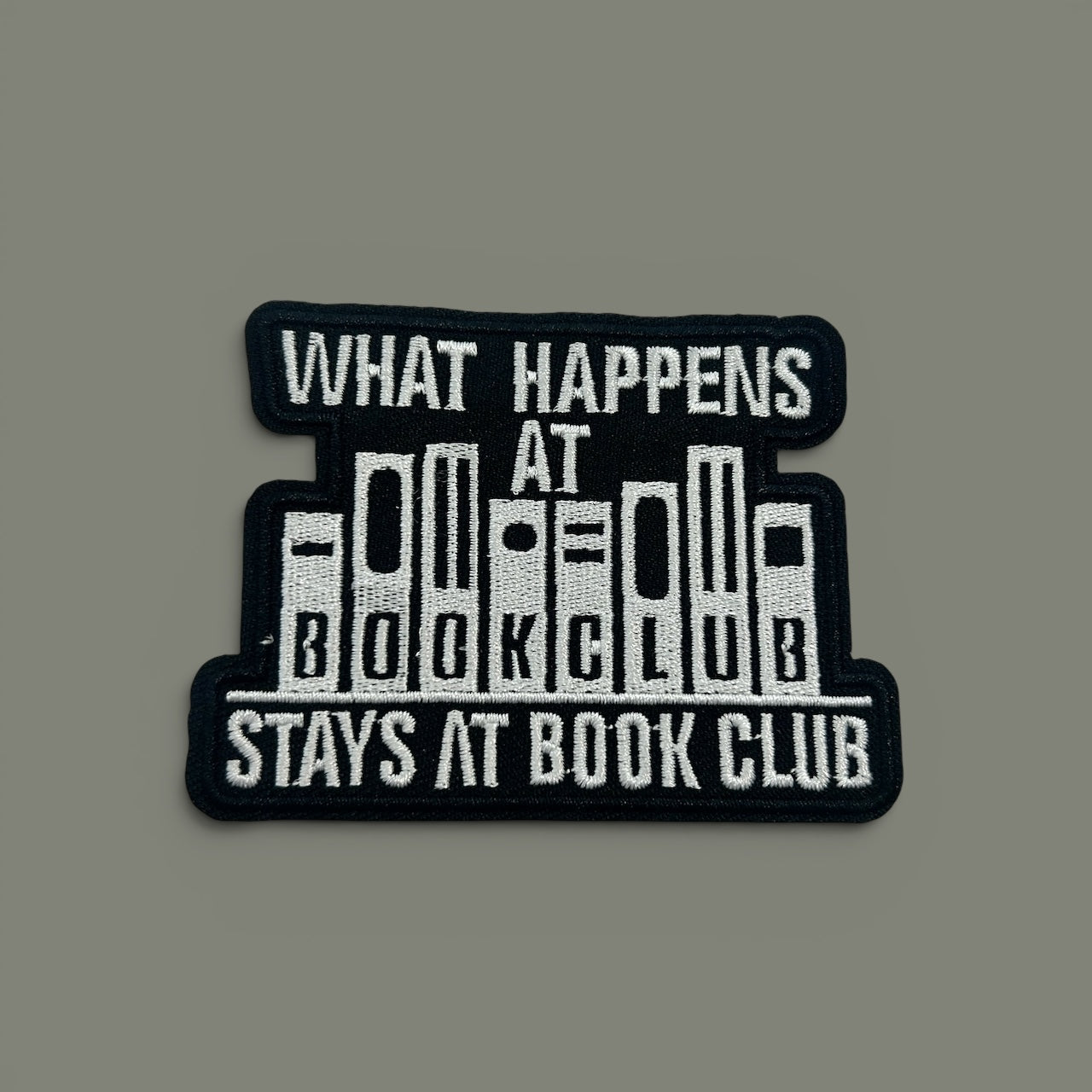 A rectangular embroidered patch with a black background and white text reading 'WHAT HAPPENS AT BOOK CLUB STAYS AT BOOK CLUB,' with stylized book spines forming the center, shown on a plain gray background.