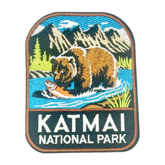 An embroidered patch featuring a brown bear catching a fish in a blue river, with mountains and trees in the background and the text 'KATMAI NATIONAL PARK' below.