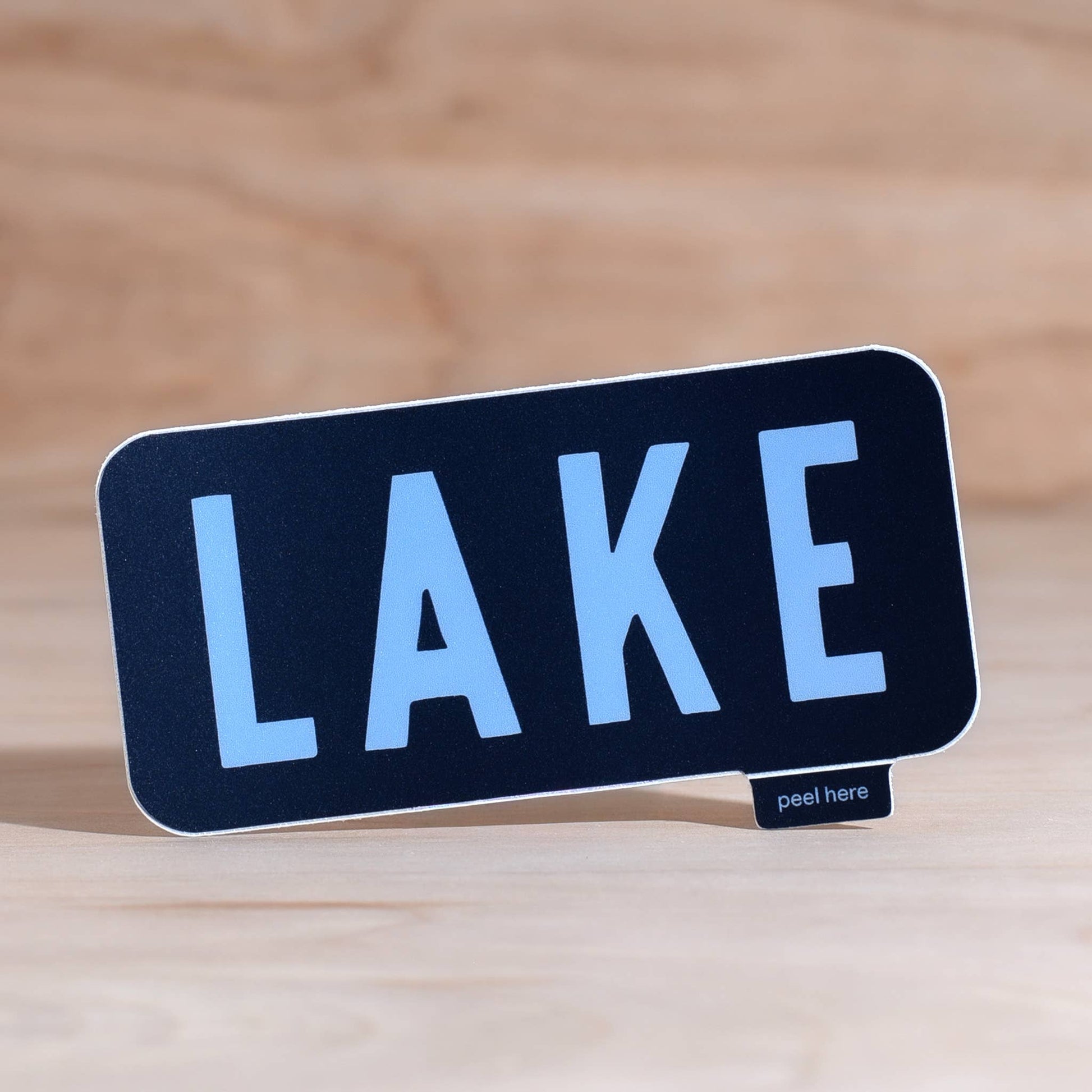 A rectangular black sign with rounded corners and the word "LAKE" in large light blue letters, shown standing on a small "peel here" tab on a light wood surface.