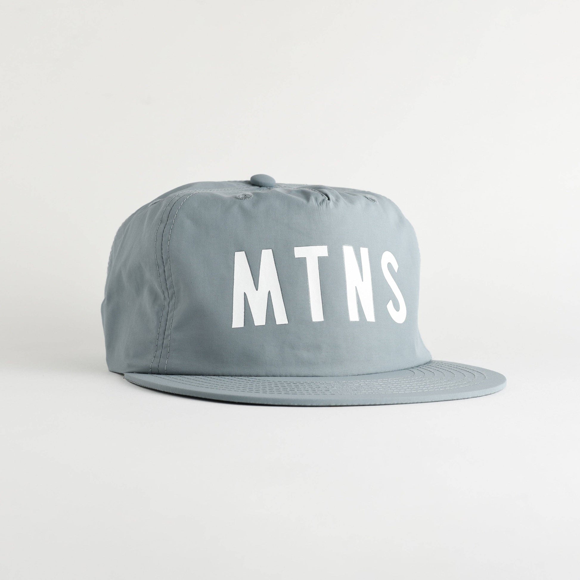 A light gray flat-brim cap with the letters 'MTNS' printed in bold white on the front, set against a plain light gray background.