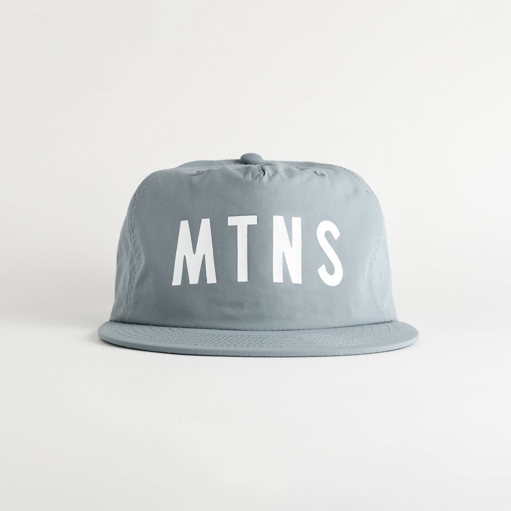 A light gray flat-brim cap with the letters 'MTNS' printed in bold white across the front, set against a plain light gray background.