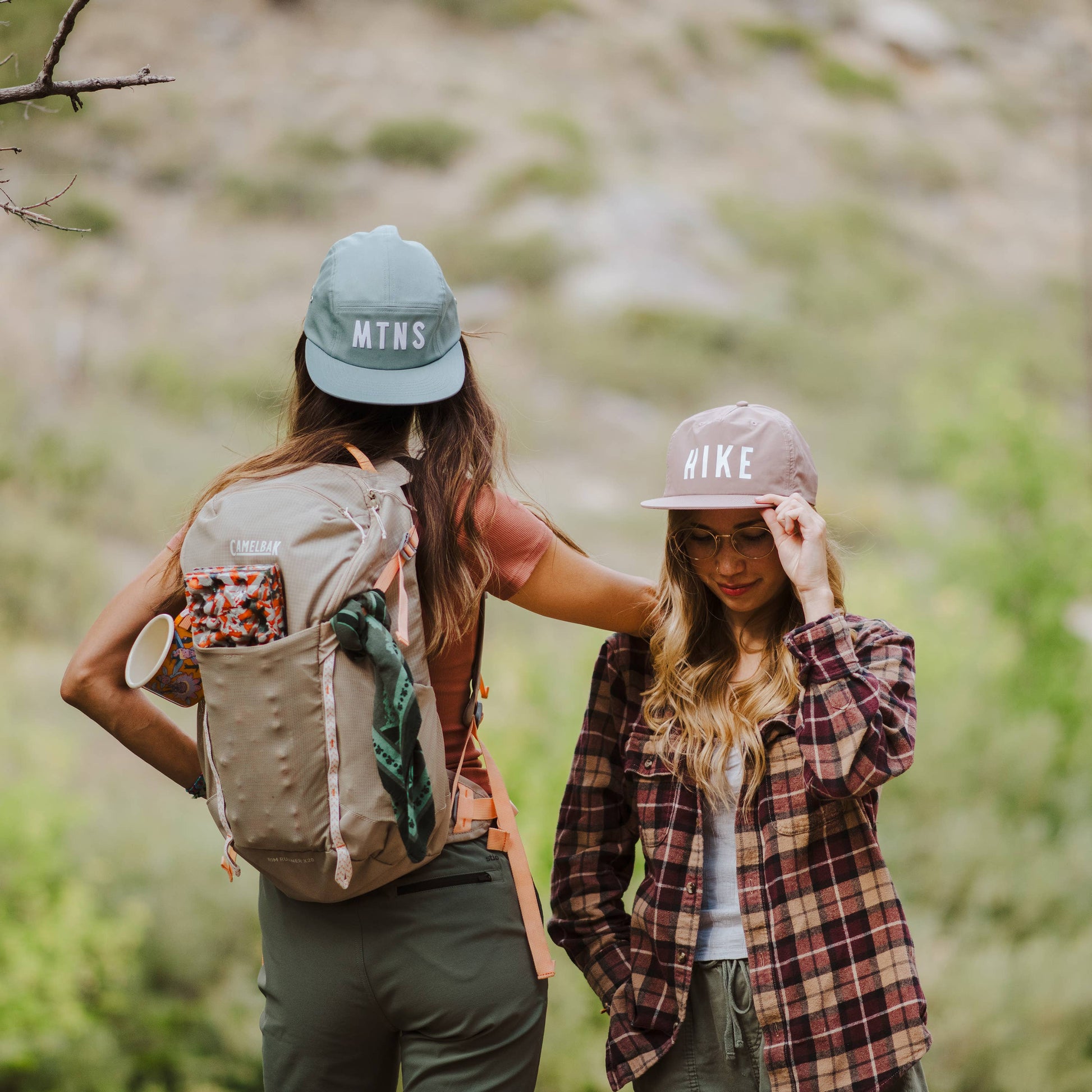 Two women hiking outdoors on a trail, seen from behind. The woman on the left wears a light blue 'MTNS' cap and a beige backpack, while the woman on the right wears a beige 'HIKE' cap and a red plaid flannel shirt.