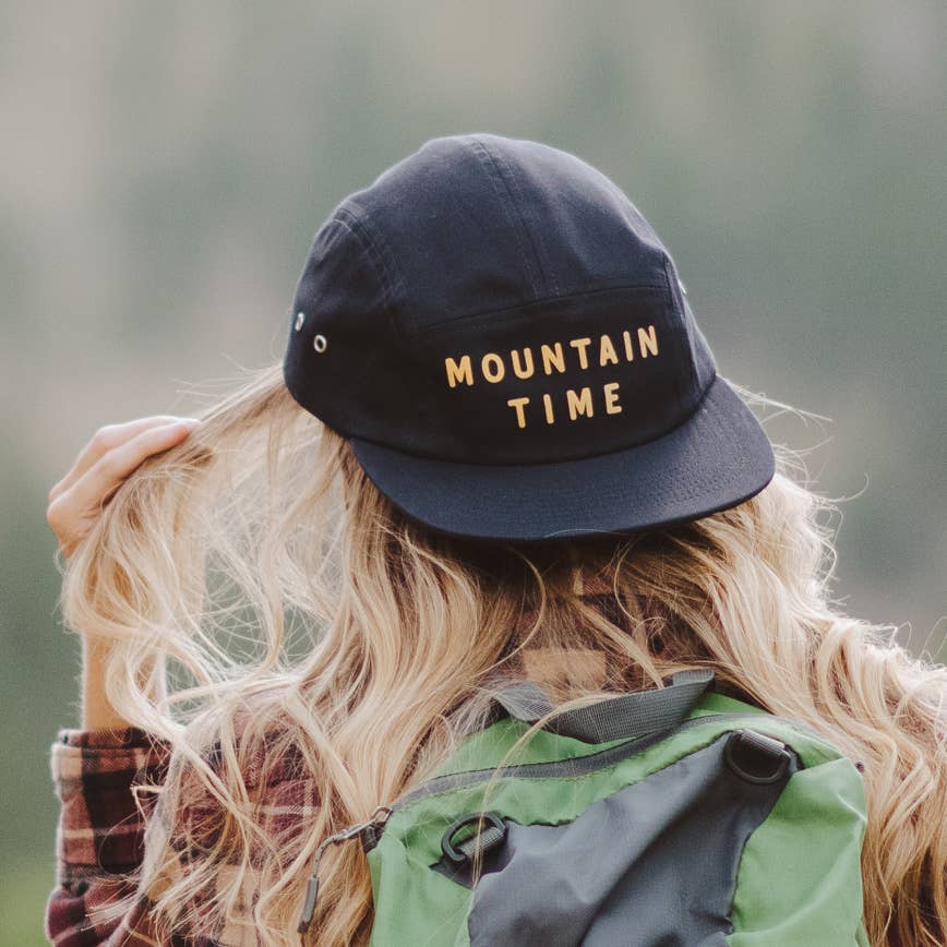 A woman seen from behind wearing a black baseball cap with 'MOUNTAIN TIME' embroidered in gold on the front, with long blonde hair and a green hiking backpack against a blurred outdoor background.