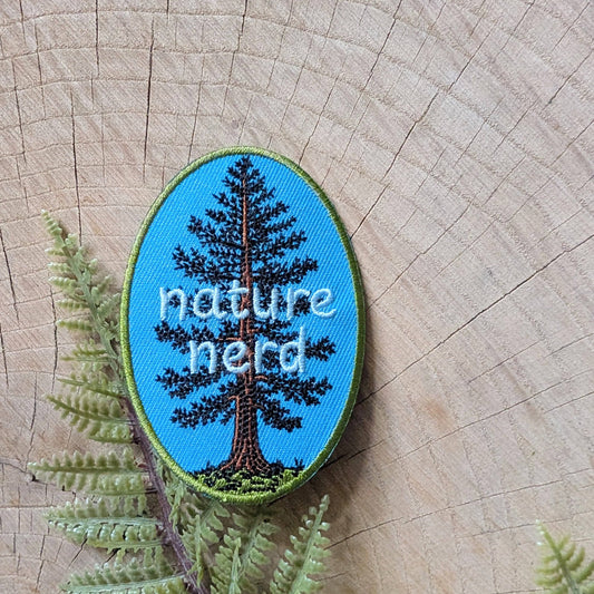 An oval embroidered patch featuring a pine tree on a bright blue background with the words 'nature nerd' in white, placed on a wood slice with green fern leaves.