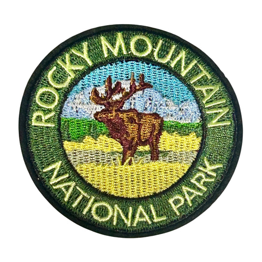 An embroidered circular patch for Rocky Mountain National Park, featuring a brown moose in a yellow field with green hills, a blue sky, and white mountains, surrounded by green border text reading 'ROCKY MOUNTAIN NATIONAL PARK'.
