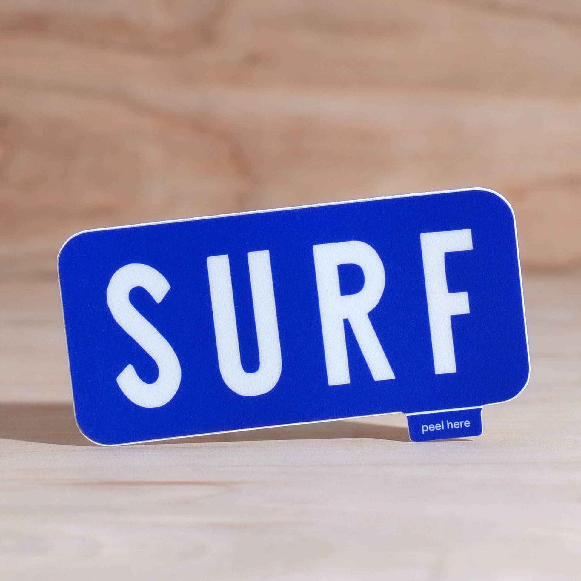 A blue rectangular sign with rounded corners and the word "SURF" in large white letters, standing on a light wood surface. A small tag at the bottom right reads "peel here.