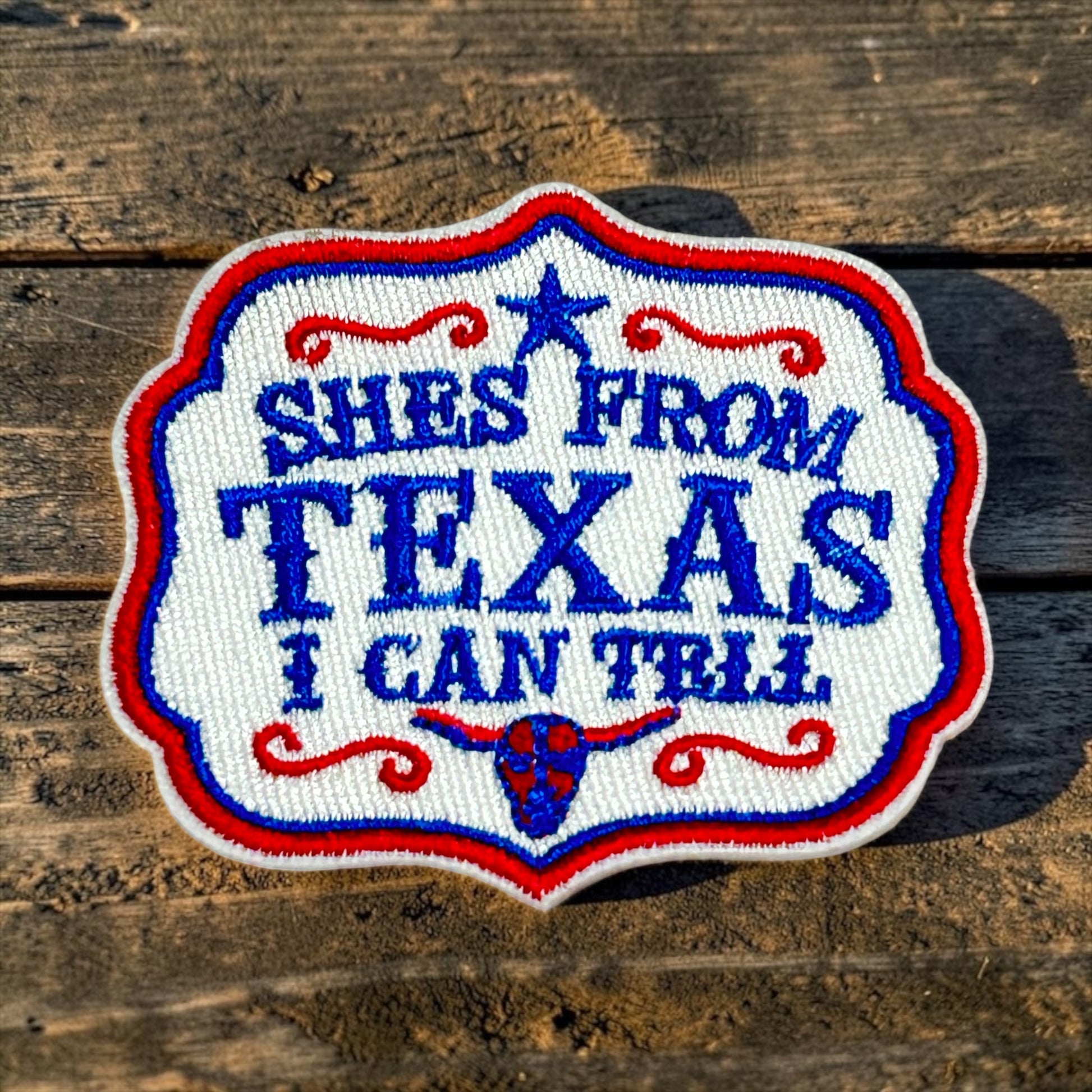 An embroidered patch with the text 'SHES FROM TEXAS I CAN TRUST' in blue lettering on a white background, bordered in red and blue, placed on a rustic wooden surface.