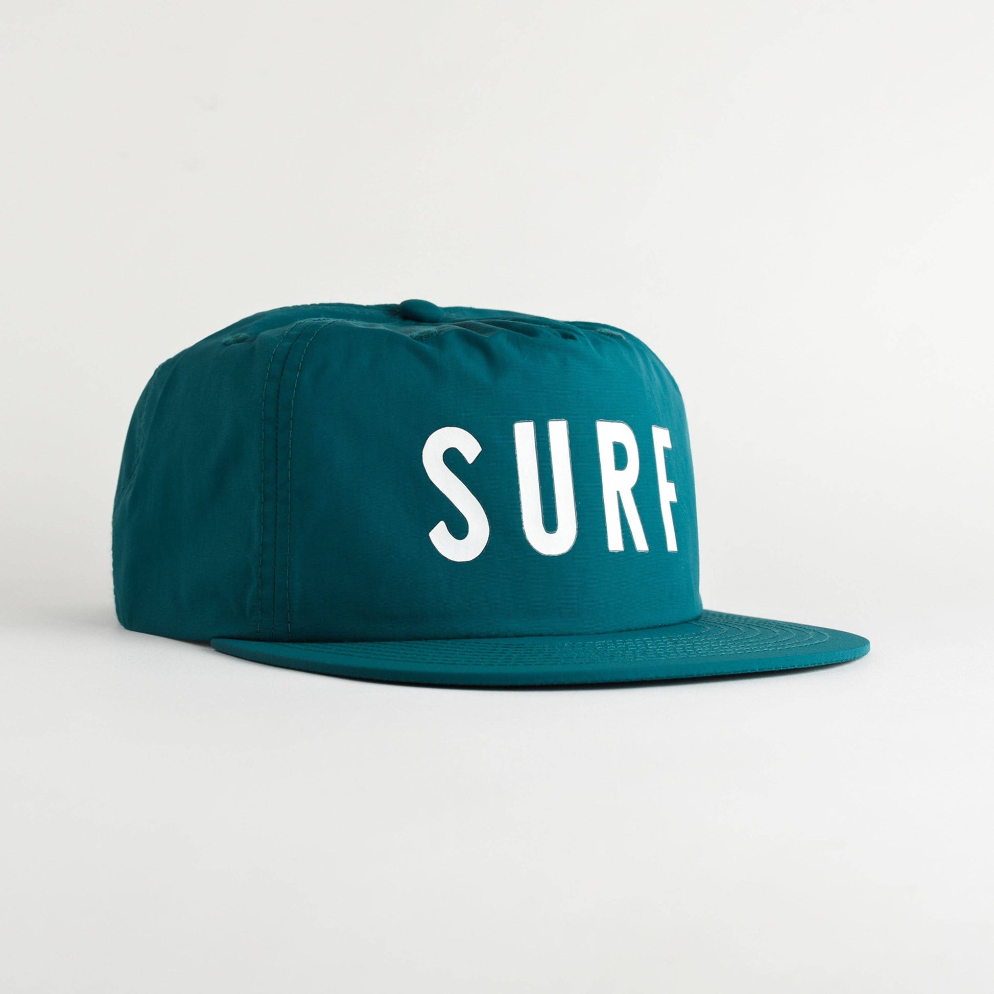 A teal snapback cap with the word 'SURF' printed in bold white letters on the front, set against a clean, light gray studio background.