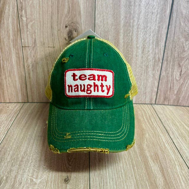 A green distressed trucker hat with mesh side panels and a white rectangular patch on the front that reads 'team naughty' in red text, photographed against a light wood plank background.