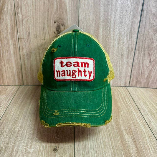 A green distressed trucker hat with mesh side panels and a white rectangular patch on the front that reads 'team naughty' in red text, photographed against a light wood plank background.